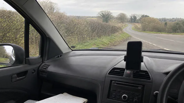 A phone mounted on a car's dashboard overlooking a winding countryside road on an overcast day, with a paper notebook on the passenger seat below