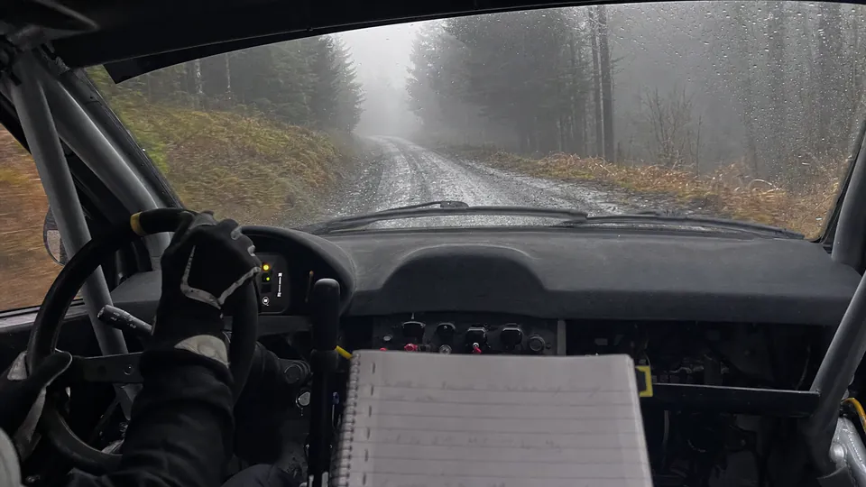 Co-driver view from a rally car on a wet, foggy forest stage with a pace-note book in the foreground